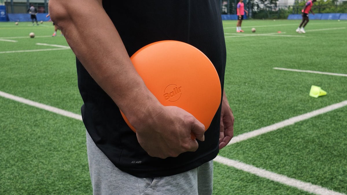 A coach carrying the Ballr AI Sports Camera under one arm beside a football training session
